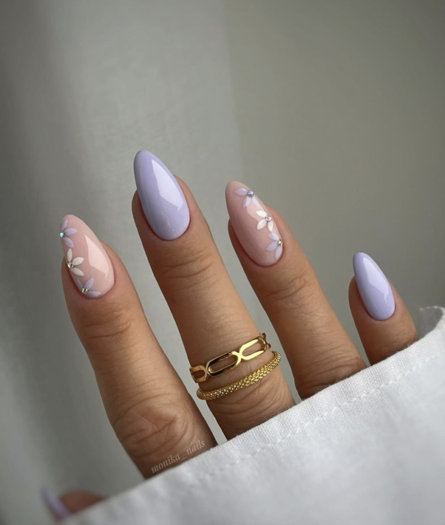 A hand models nails alternating between a solid, tender lavender color and a nude base with sophisticated white floral art and subtle rhinestone embellishments.